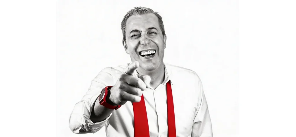 Hombre sonriente con corbata roja, señalando hacia la cámara.